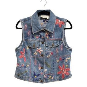 May include: A blue denim vest featuring a button-front closure, a pointed collar, and two chest pockets. The garment is decorated with colorful floral and leaf embroidery patterns, along with decorative stitching lines across the front panels.