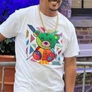 May include: A white short-sleeved t-shirt featuring a central graphic of a multi-colored teddy bear with X-shaped eyes. The bear is surrounded by various geometric shapes in shades of blue, pink, and yellow. A gold-toned chain necklace is worn.