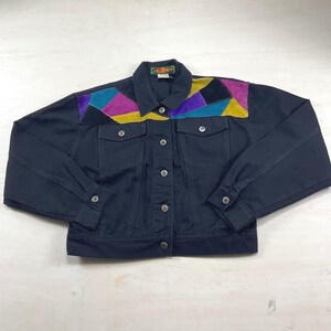 May include: A black denim jacket featuring a multi-colored geometric patch design across the upper chest and shoulders in shades of teal, magenta, purple, and yellow. The jacket includes a button-front closure, two chest pockets, and a collar.