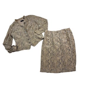 May include: A two-piece set consisting of a long-sleeved button-down jacket and a matching knee-length skirt. Both pieces feature a tan and black snakeskin print pattern. The jacket has a collared neckline and a row of dark buttons down the front.