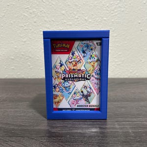 May include: A blue, 3D-printed frame holds a Pokemon Prismatic Evolutions Booster Bundle. The bundle features colorful diamond-shaped images of Pokemon characters. The frame sits on a gray wooden surface.