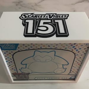 Pode incluir: Uma vitrine retangular branca contendo uma caixa de treinador de elite Pokémon Scarlet & Violet 151. O topo da caixa apresenta o texto Scarlet & Violet 151 em preto e branco. A caixa interna exibe um gráfico do Snorlax e ícones coloridos.
