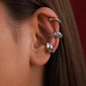 May include: Three silver ear cuffs are shown on an ear. The top cuff is a simple curved bar. The middle cuff has a textured design, possibly with gems. The bottom cuff is a smooth, rounded shape.