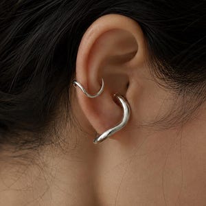Peut inclure: Deux bagues d'oreille argentées au design lisse, ondulé et incurvé. La plus petite bague est placée sur le cartilage supérieur, tandis que la plus grande et plus épaisse repose sur la partie médiane de l'oreille, créant un aspect métallique superposé.