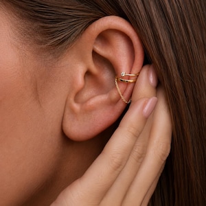 Puede incluir: Puño de oreja dorado con una pequeña gema transparente en la banda superior. Una delicada cadena dorada conecta las bandas superior e inferior. La joya se lleva en la oreja.