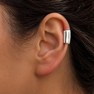 Pode incluir: Um ear cuff prateado é usado na parte superior da orelha. O ear cuff tem uma forma retangular sólida com uma superfície lisa e reflexiva. O ear cuff é uma joia moderna.