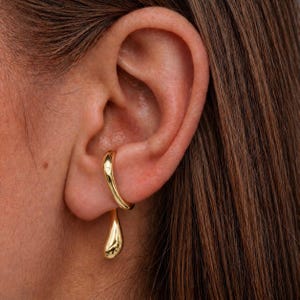 Könnte beinhalten: Ein goldfarbenes Ohrring-Set. Der Ohrring hat ein zweiteiliges Design, mit einem gebogenen oberen Teil, der sich um das Ohr legt, und einem tropfenförmigen Anhänger. Der Schmuck ist glänzend und reflektierend, mit einer glatten Oberfläche.