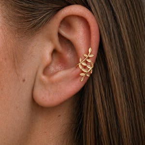 Puede incluir: Un ear cuff dorado con un diseño de hojas. El brazalete se envuelve alrededor de la oreja, con múltiples hojas dispuestas en un patrón decorativo. Esta joya añade un toque de elegancia inspirada en la naturaleza.
