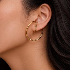Può includere: Un paio di orecchini a cerchio color oro e un ear cuff più piccolo sono indossati sull'orecchio. Il grande orecchino a cerchio è circolare e l'ear cuff è una fascia curva più piccola che si posiziona sulla parte superiore dell'orecchio. I gioielli sono lucenti.