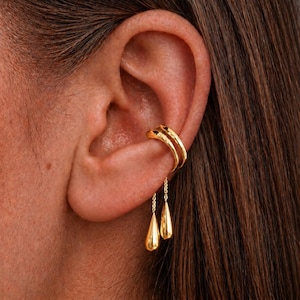May include: A gold ear cuff and drop earrings are shown close up. The ear cuff has a double-band design and wraps around the ear cartilage. Two teardrop-shaped earrings dangle from fine chains, creating an elegant and stylish appearance.