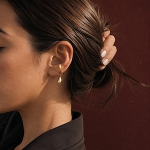 May include: A close-up of a gold-colored earring set. The earring set includes a hoop earring and a teardrop-shaped dangle earring. The earrings are worn on the ear of a person with dark hair and a black top. The background is a solid maroon color.