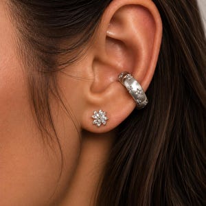 May include: A silver ear cuff and a matching stud earring set. The ear cuff features a star pattern, while the stud earring is a cluster of small, sparkling stones. The jewelry is worn on an ear.
