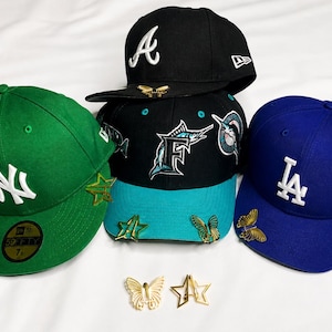 Puede incluir: Gorras de béisbol en negro, verde y azul, cada una adornada con logotipos de equipos y alfileres decorativos. Las gorras presentan logotipos bordados en blanco y alfileres de mariposa y estrella doradas.
