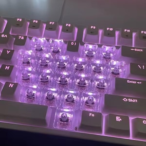 Könnte beinhalten: Nahaufnahme einer weißen mechanischen Tastatur mit transparenten Tastenkappen, die von violetten LEDs beleuchtet werden. Die Tastenkappen sind in Herzform angeordnet, was ein einzigartiges Design ergibt. Die Tastatur hat Standard-Funktionstasten.