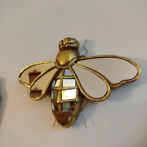 以下が含まれることがあります： 金色のフレームと鏡張りの羽を持つ、蜂の形をした装飾的な壁掛け鏡。 蜂の体は小さな鏡張りの正方形で区切られています。 蜂には2本の触角があり、白い壁に取り付けられています。