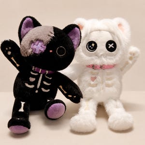 Pode incluir: Dois brinquedos de pelúcia: um gato preto com detalhes de esqueleto e um adesivo de cruz roxa, e um urso branco em uma roupa com tema de esqueleto. Ambos têm coleiras e um design gótico assustador.