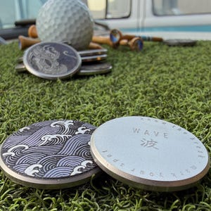 May include: Two circular metal golf ball markers resting on green artificial turf. One marker features a purple and black wave pattern, while the other is silver with the text WAVE, the Japanese character for wave, and Know the break.