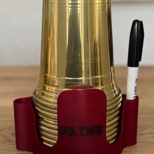 May include: A stack of gold-colored cups in a burgundy holder with the name "PAINE" printed on the front. A black and white marker is placed in a side slot. The cups and holder are on a wooden surface.
