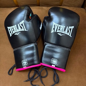 Può includere: Un paio di guantoni da boxe neri con il logo bianco Everlast sulla parte anteriore. Ogni guanto presenta un'etichetta con la scritta Hecho en Mexico e un bordo rosa acceso sul polso. I guantoni sono dotati di lacci neri per la chiusura.