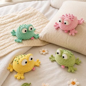 May include: Four crocheted frog toys in shades of green, yellow, and pink, with large googly eyes and small pink tongues. The frogs are lying on a bed with a cream-colored blanket and pillows, with a few crocheted flower accents.