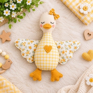 Könnte beinhalten: Ein Enten-Stofftier mit cremefarbenem Kopf und gelber Vichy-Karo-Schleife, einem Körper mit gelbem Vichy-Karo-Muster und Herz-Applikation sowie Flügeln mit kleinen gelben Blumen. Die Ente hat orangefarbene Füße, einen gelben Schnabel und rosa Wangen.