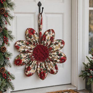 May include: A fabric flower-shaped wall hanging featuring a cream background with a red and green poinsettia and holly leaf pattern. The center is a dark red woven fabric, and the piece includes a matching fabric loop for hanging on a hook.