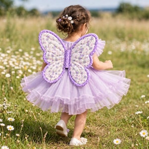 Puede incluir: Un vestido color lavanda con falda de tul de varias capas y mangas con volantes, combinado con alas de mariposa acolchadas con borde de lunares morados y blancos y centro de estampado floral, usado con zapatos blancos y accesorios florales.