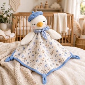 Könnte beinhalten: Eine Kuscheldecke mit einem weißen Entenkopf, gelbem Schnabel, geschlossenen Augen und rosa Wangen. Die Ente trägt ein blaues Barett und ein blau-weiß kariertes Halstuch. Der Körper der Decke hat ein blaues Blumenmuster und verknotete Ecken.