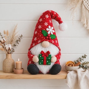 Può includere: Una statuetta di gnomo in peluche che indossa un alto cappello rosso con motivo a fiocchi di neve bianchi, un pompon bianco e un dettaglio in feltro a forma di agrifoglio. Lo gnomo ha una lunga barba bianca, naso tondo e tiene un regalo verde.