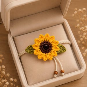 May include: A handmade crochet bracelet featuring a yellow sunflower with a brown center and green leaves. The bracelet is displayed in an open beige jewelry box. The bracelet has a cream-colored band with wooden beads.