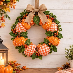 Pode incluir: Uma guirlanda decorativa com abóboras de tecido em padrões xadrez e florais laranja e branco, acentuada com folhas de feltro verde, pequenas bagas laranja e caules castanhos. Um grande laço de juta está atado no topo para pendurar.