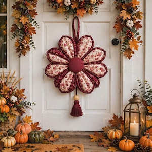 Könnte beinhalten: Ein Stoffblumen-Wandbehang mit acht gesteppten Blütenblättern in einem dunkelroten und cremefarbenen Blumenmuster. Die Mitte ist ein einfarbiger dunkelroter Kreis. Eine Holzperle und eine passende dunkelrote Quaste hängen unten.
