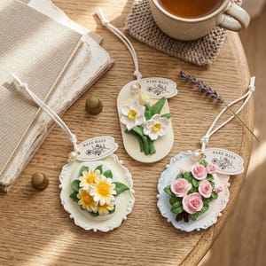 May include: Three handmade floral ornaments with white string, each featuring a different flower arrangement: daffodils, daisies, and roses. Each ornament has a tag that says "HAND MADE". The ornaments are on a wooden table.