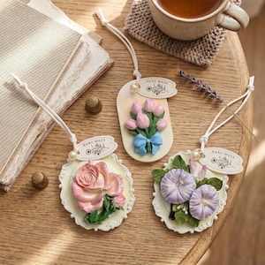 May include: Three oval-shaped hanging ornaments featuring relief floral designs: a pink rose, a bouquet of pink tulips with a blue bow, and purple morning glory flowers. Each piece includes a white cord, a pearl bead, and a tag labeled HAND MADE.
