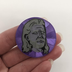 May include: A purple and gray 3D printed pin featuring a portrait of a man with long hair. The pin has a textured, circular design.