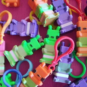 May include: A collection of colorful, 3D-printed keychains in various colors, including orange, green, purple, and pink. Each keychain features a unique, blocky design with a clip for attachment. The keychains are scattered on a pink surface.