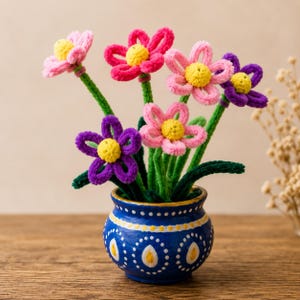 Può includere: Un bouquet di sei fiori all'uncinetto con petali rosa, magenta e viola e centri gialli, ciascuno su uno stelo verde. I fiori sono disposti in un piccolo vaso di ceramica blu decorato con punti bianchi e motivi a goccia di colore giallo.