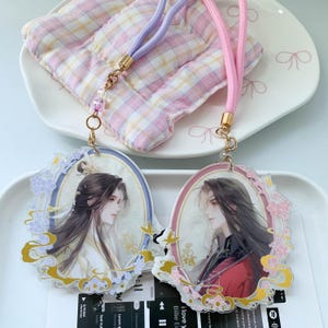May include: Two oval acrylic charms featuring anime-style illustrations of characters with long dark hair, framed by decorative floral borders in purple and pink. Each charm is attached to a matching pastel-coloured cord with gold-tone metal clasps.