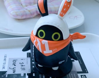 May include: A stylised black figurine with large yellow eyes, wearing a white hood with rabbit ears, an orange bandana with the white text 1 and EQUUS, and a black jacket. The figure stands on a circular base, featuring a small white tag with the text i still love you.