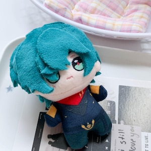 Peut inclure: Une poupée en peluche avec des cheveux bleu sarcelle, des yeux brodés aux iris verts et un teint pâle. La poupée porte une tenue bleu foncé avec des accents jaunes et un col rouge. La peluche a une texture douce et une forme de tête arrondie.
