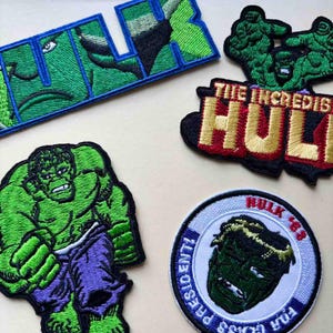 Könnte beinhalten: Gestickte Aufnäher mit Hulk-Motiven. Die Aufnäher zeigen das Wort "HULK" in Grün und Blau, eine grüne Hulk-Figur und den Schriftzug "THE INCREDIBLE HULK" in Rot und Gold. Ein Aufnäher hat den Text "HULK '63".