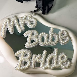 Puede incluir: Letras blancas cubiertas de perlas que deletrean "Mrs Babe Bride" sobre una superficie espejada. Las letras están delineadas con pequeños diamantes de imitación transparentes. El arreglo se encuentra sobre una bandeja blanca y curvada, con una rosa negra en el fondo.