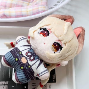 Peut inclure: Une poupée en peluche représentant un personnage aux cheveux blond clair, aux oreilles pointues et aux grands yeux brodés. La poupée porte une chemise blanche avec des détails à motifs bleus et noirs, un nœud rouge et des ornements verts.