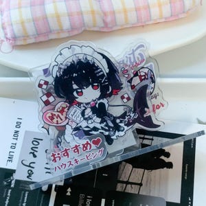 May include: A clear acrylic standee featuring a chibi-style character with black hair and red eyes, wearing a black and white maid outfit. The character has a shark tail and is surrounded by red and white checkered patterns. Text in Japanese reads "おすすめ♡ハウステーピング".
