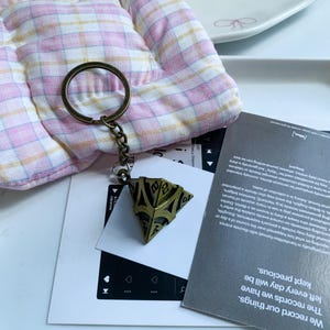 May include: A metal keychain featuring a bronze-toned, multi-faceted pyramid charm with engraved numbers on each triangular face. The charm is attached to a circular metal ring by a short chain, displaying a weathered, antique-style finish.