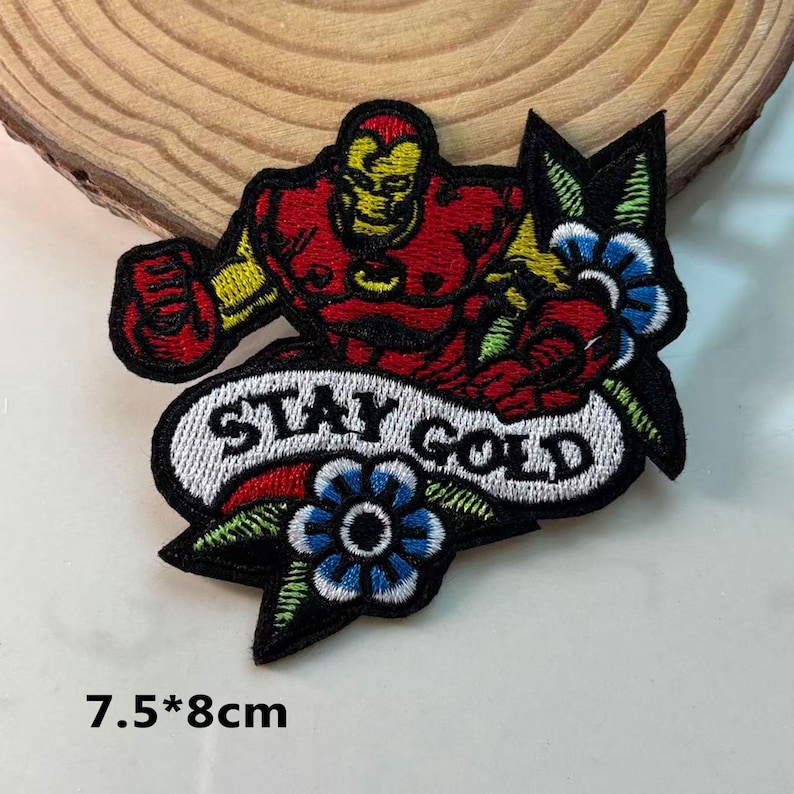 May include: Embroidered patch featuring a red and yellow Iron Man design with the words "STAY GOLD" on a white banner. The patch includes blue flowers and measures 7.5 x 8 cm.