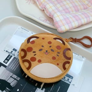 May include: A round, plush, tan coin purse featuring a stylized leopard face with dark brown spots, a cream-colored belly patch, and a small brown plastic clip attached to the side zipper. The purse has embroidered facial features.
