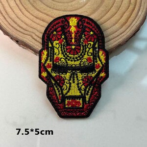 May include: Embroidered patch in the shape of a stylized face, featuring a red, yellow, and black colour scheme. The design incorporates floral and geometric patterns. The patch measures 7.5*5cm.
