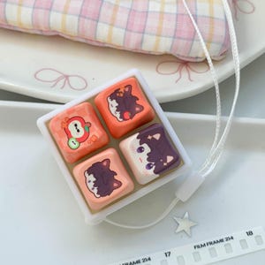May include: A square white tray containing four mechanical keyboard keycaps with colorful cartoon illustrations of characters and an apple, attached to a white lanyard with a small silver star charm and a ruler showing 17 to 18 inches.