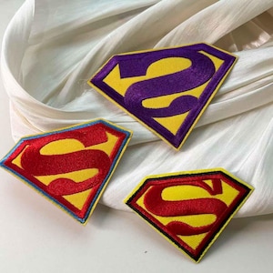 Pode incluir: Três patches bordados em forma de diamante com fundo amarelo e uma grande letra "S" em vermelho, roxo e preto. Os patches têm uma borda amarela. Os patches estão em uma superfície branca texturizada.